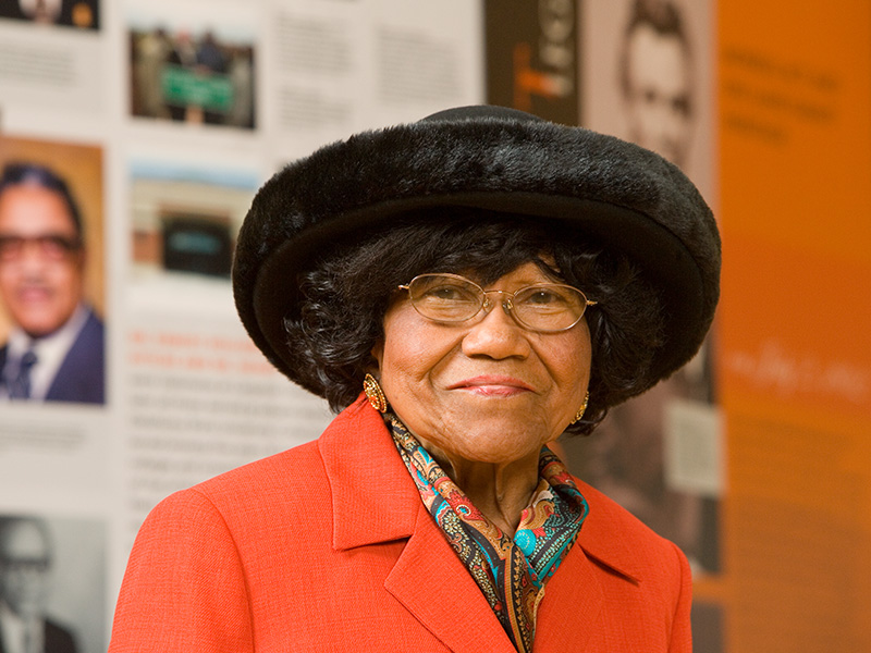 Nancy Randolph Davis, a Black woman wearing an orange blazer and a black hat, stands in an OSU Hallway